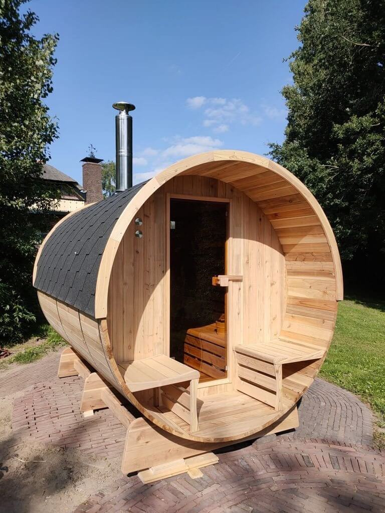 Ronde barrelsauna van red ceder hout. Met glazen deur en houtkachel, vooraan de sauna ook twee zitjes.